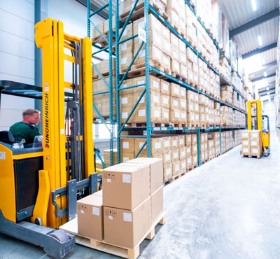 WKK Mitarbeiter zwischen zwei Hochregalen in der WKK Lagerhalle. Mit zwei Gabelstaplern mit jeweils einer Palette und Kartons.