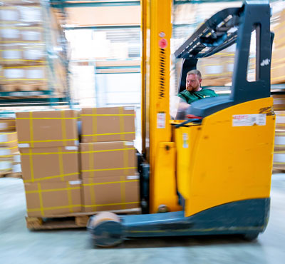 Ein WKK Mitarbeiter im Lager, der auf einem Gabelstapler eine Palette mit Kartons transportiert. Aufgenommen während der Fahrt.