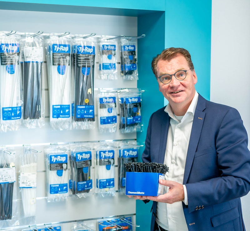 Ein WKK Mitarbeiter steht vor einem Ty-Rap Regal und hält eine Arbeitsbox mit schwarzen Kabelbindern der Marke Ty-Rap von der Firma ABB Thomas & Betts.