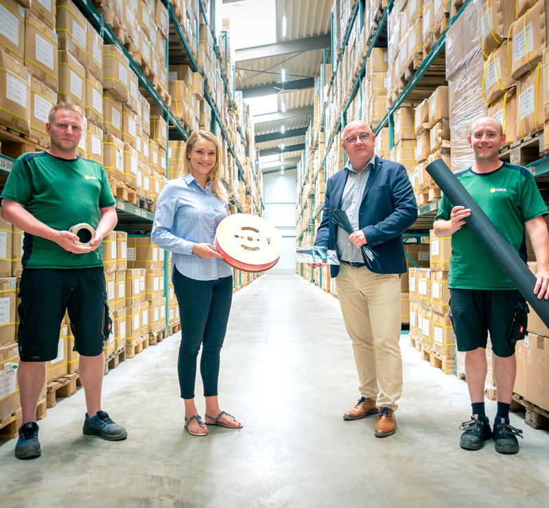 Vier WKK Mitarbeiter im Lager halten jeweils ein Produkt verschiedener Produktkategorien. Eine Kabelverschraubung aus Metall, eine rote Schrumpfschlauchrolle, ein Bündel schwarzer Kabelbinder und die WKK Kabelbinderzange sowie eine schwarze Schrumpfschlauchstange von 1220mm Länge.